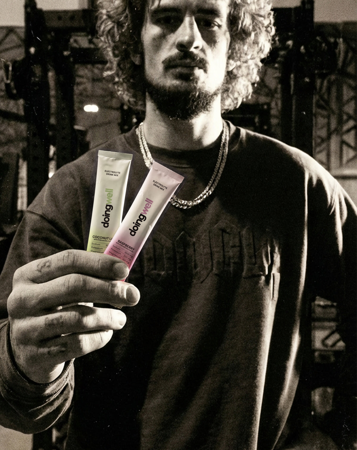 Black and white gym photo of male athlete with curly hair wearing a sweatshirt and chain necklace, holding both DoingWell stick packs in color—Coconut Lime (green) and Raspberry (pink)—with gym equipment visible in the background