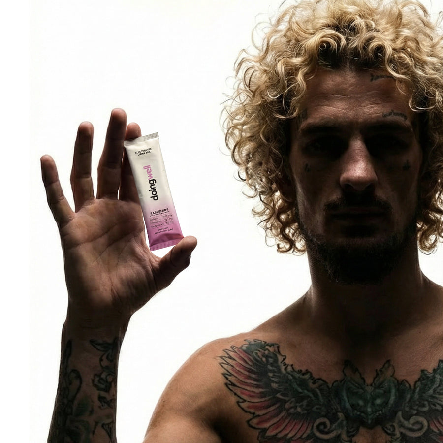 Sean O'Malley shirtless with curly blonde hair and signature owl wing chest tattoo, holding a DoingWell Raspberry stick pack between his fingers with arm extended, face tattoo visible, against a white background