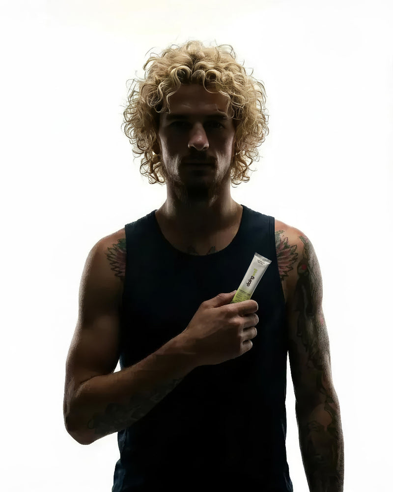 Sean O'Malley in a black tank top holding a DoingWell Coconut Lime stick pack against his chest, curly blonde hair backlit against a bright white background, with colorful arm tattoos visible