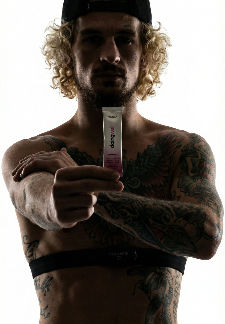 Sean O'Malley with curly blonde hair and black backwards cap, shirtless with extensive arm and torso tattoos, hand wraps and heart rate monitor strap visible, holding a DoingWell Raspberry stick pack toward the camera, dramatic backlit silhouette lighting against white background